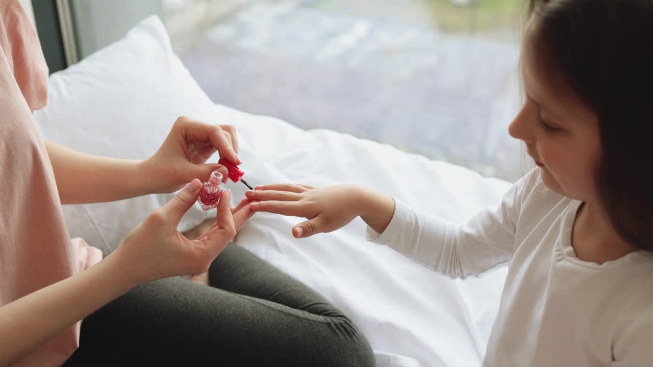 年轻快乐的女人坐在床上给她的小女孩涂指甲。视频下载