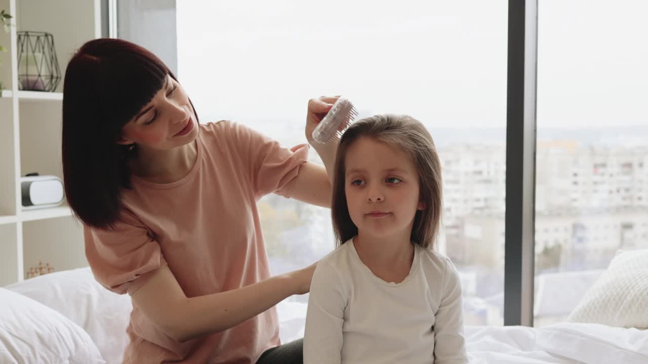 可爱的小女孩微笑着，她快乐的妈妈正在给女儿梳头。视频下载