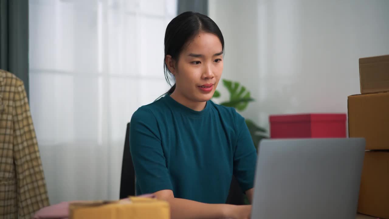 年轻的女企业主坐在办公桌前使用笔记本电脑，同时检查订单和写在书上。视频下载