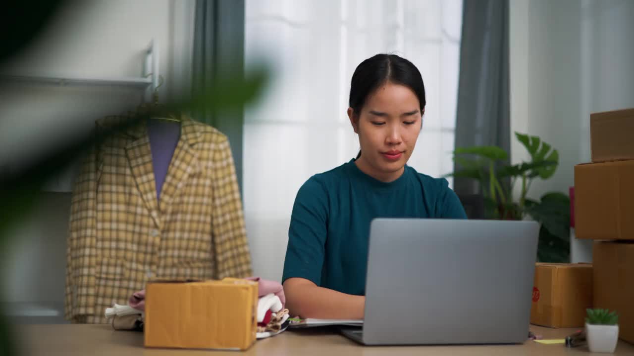 年轻的女企业主坐在办公桌前使用笔记本电脑，同时检查订单和写在书上。视频下载