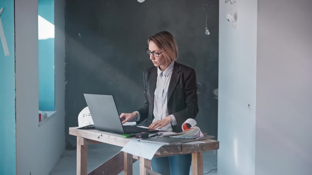 一名身着正装的女子站在办公桌旁，手里拿着笔记本电脑和文件视频下载