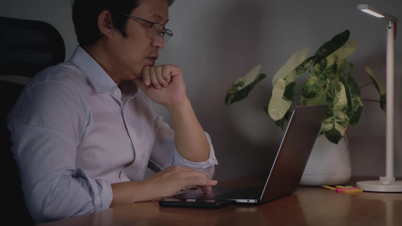一个男人坐在桌子前，手里拿着一台笔记本电脑和一盆植物。他在笔记本电脑上打字，陷入了沉思。当一个人在电脑上工作时，注意力集中的概念视频下载