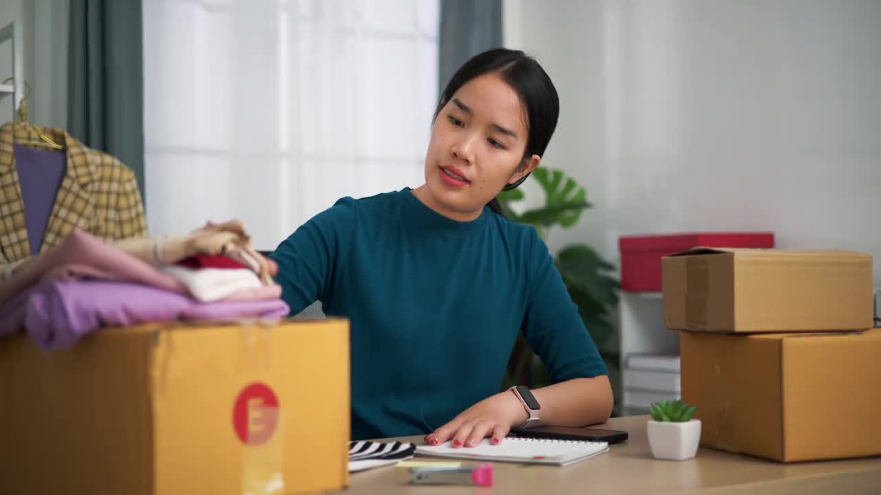年轻的女企业主坐在办公桌前检查订单，用智能手机扫描箱子包装上的二维码。视频下载