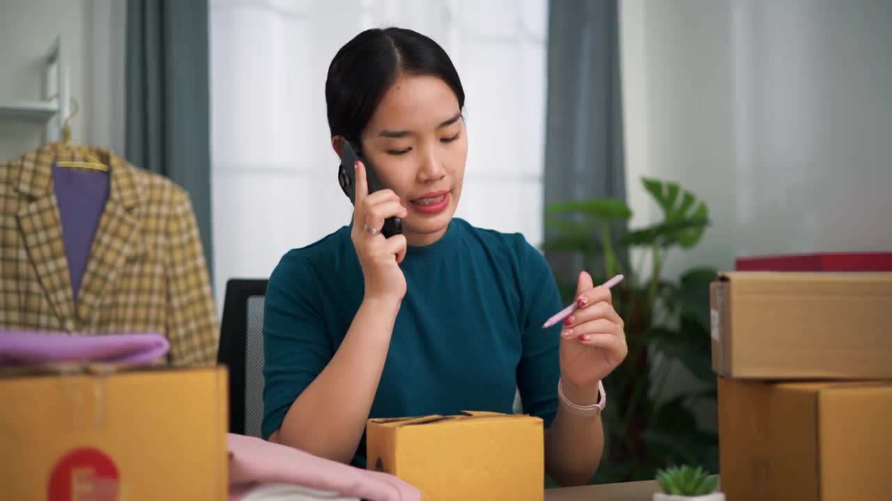年轻的女企业主坐在办公桌前用智能手机与客户交谈，同时打包运输。视频下载