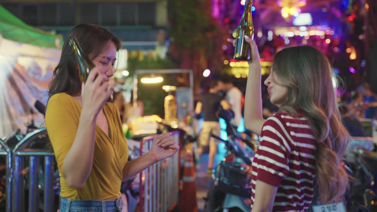 亚洲女性朋友捧着一瓶啤酒，在酒吧前聚会。美丽的两位女旅行者在城市户外旅行，享受在夜总会度过庆祝节日的假期旅行。视频下载
