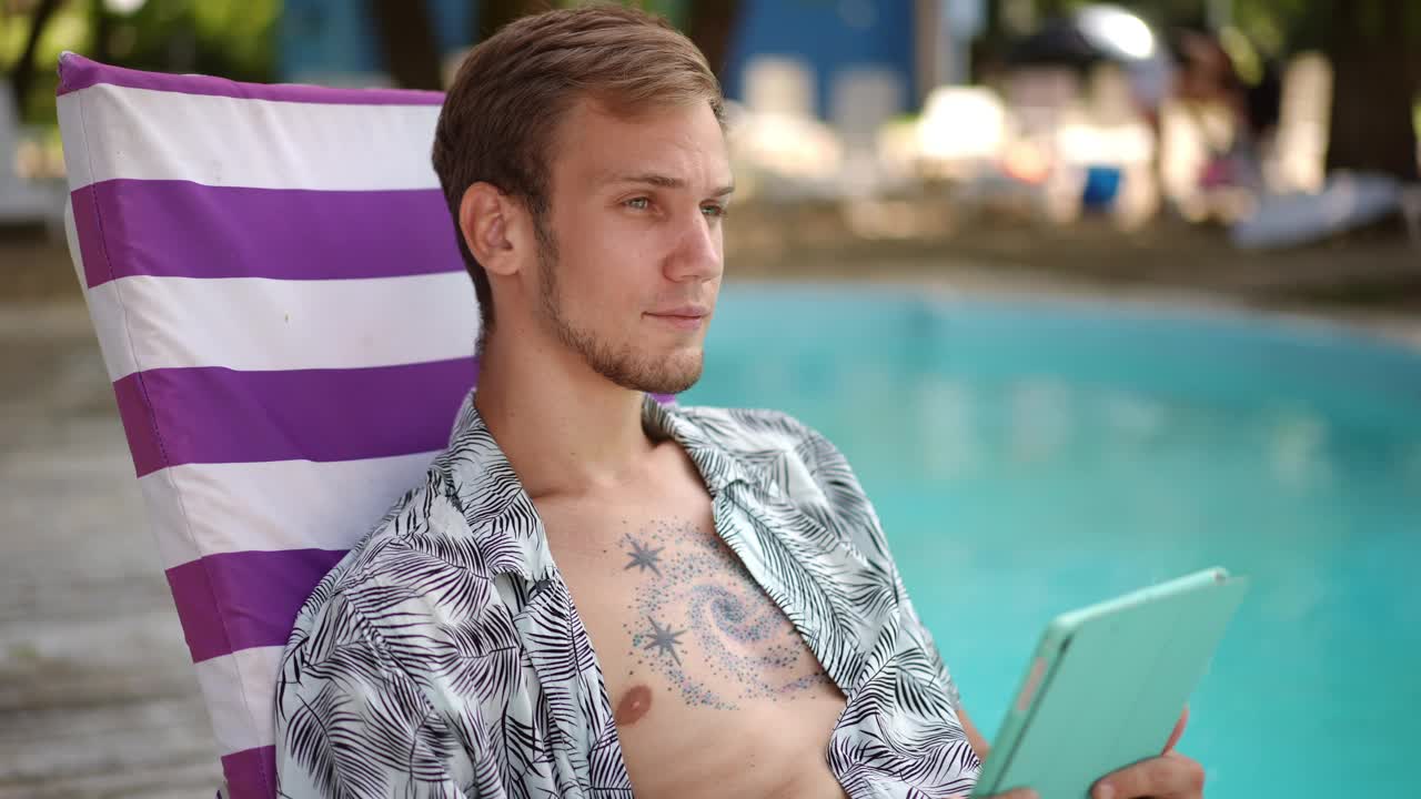缓慢的运动。特写镜头。在一个阳光明媚的夏日，一个年轻人坐在沙滩椅上，手里拿着平板电脑，在游泳池附近摆姿势。这个人笑着看着镜头。视频下载