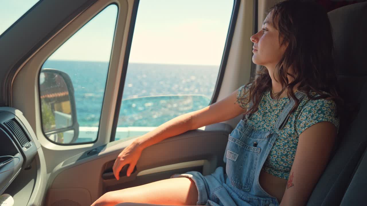 一名妇女坐在露营车的副驾驶座位上，看着窗外的海边视频下载