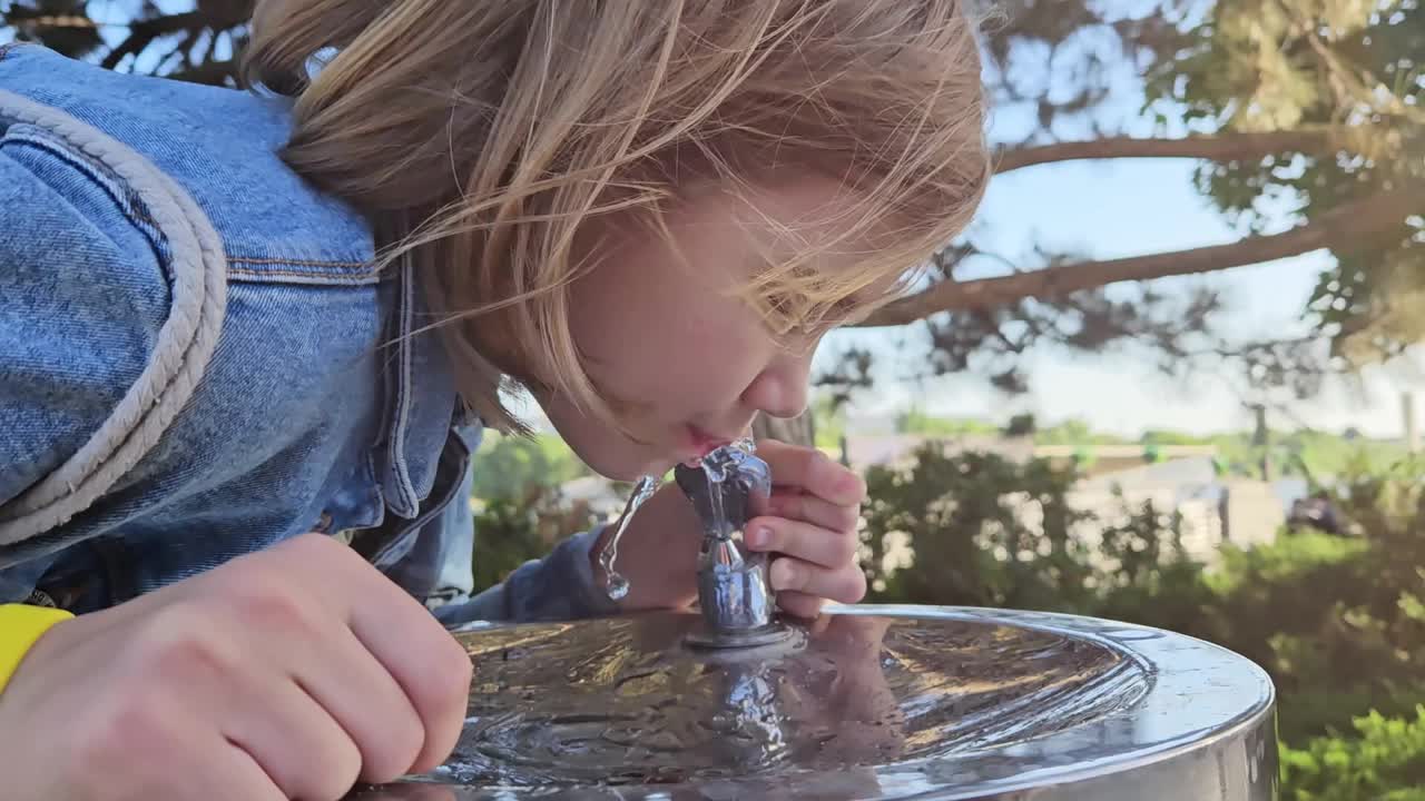 穿着牛仔夹克的少女在喷泉喝水。视频下载