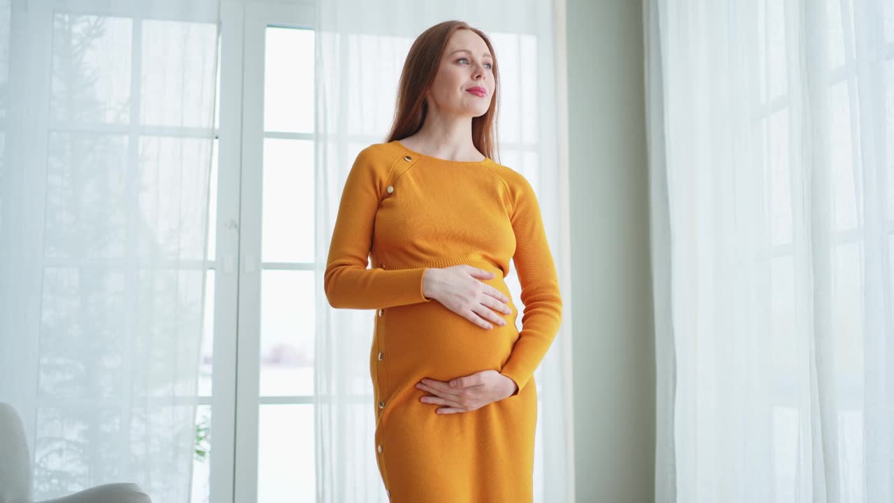 怀孕做母亲的人对未来的期望。大肚子的孕妇站在家里的窗户旁边。女孩抱着肚子享受怀孕。母性柔情亲子新生活理念。视频下载