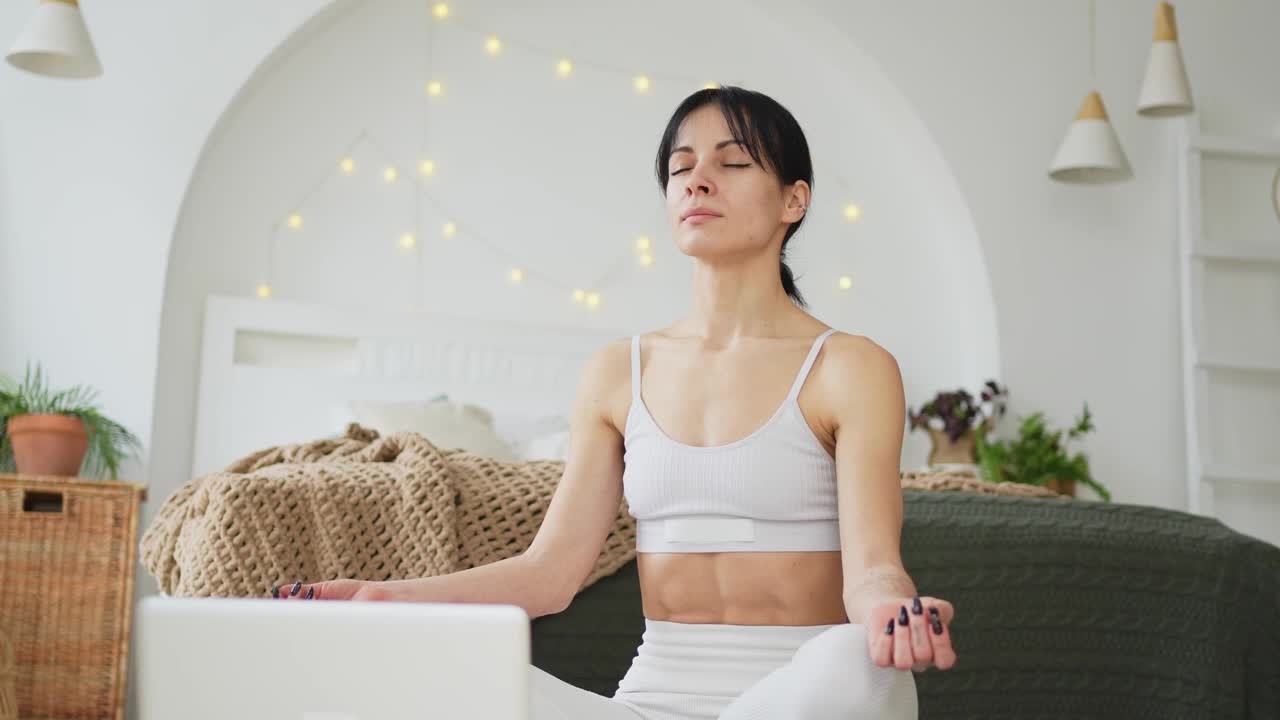 瑜伽正念冥想在线。妇女在家用笔记本电脑练习在线瑜伽课程。坐在莲座里的女人在室内冥想放松。女孩在做呼吸练习。在家做瑜伽。视频下载