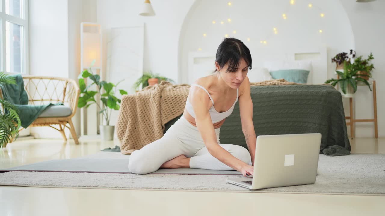 在线瑜伽健身锻炼训练。妇女在家用笔记本电脑练习在线瑜伽课程。在瑜伽垫上做运动的妇女。正在进行减肥训练的女孩。运动健身。视频下载