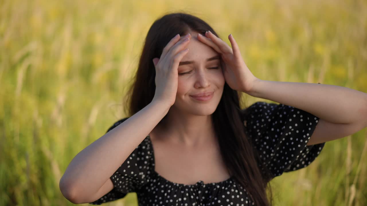 一个可爱的黑发女孩闭着眼睛，把头转向太阳，微笑着，站在夏日的田野里，背景是黄色的草地。视频下载