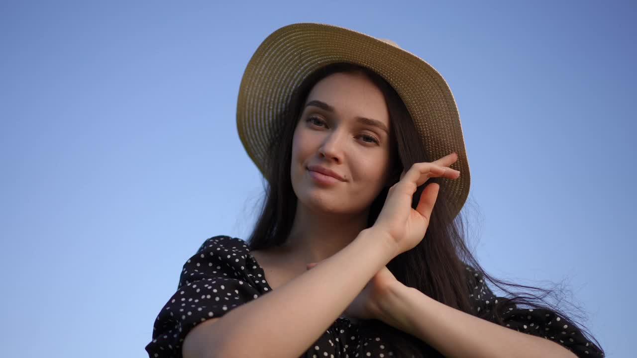 在夏日蔚蓝的天空背景下，一个戴着帽檐帽的黑发女孩摆姿势，微笑着看着镜头。低角。视频下载