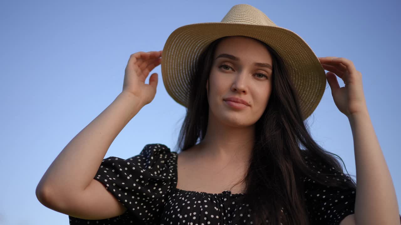 美丽的黑发年轻女子戴着一顶有檐的帽子，站在夏日户外的蓝天下，看着镜头微笑着。低角。视频下载