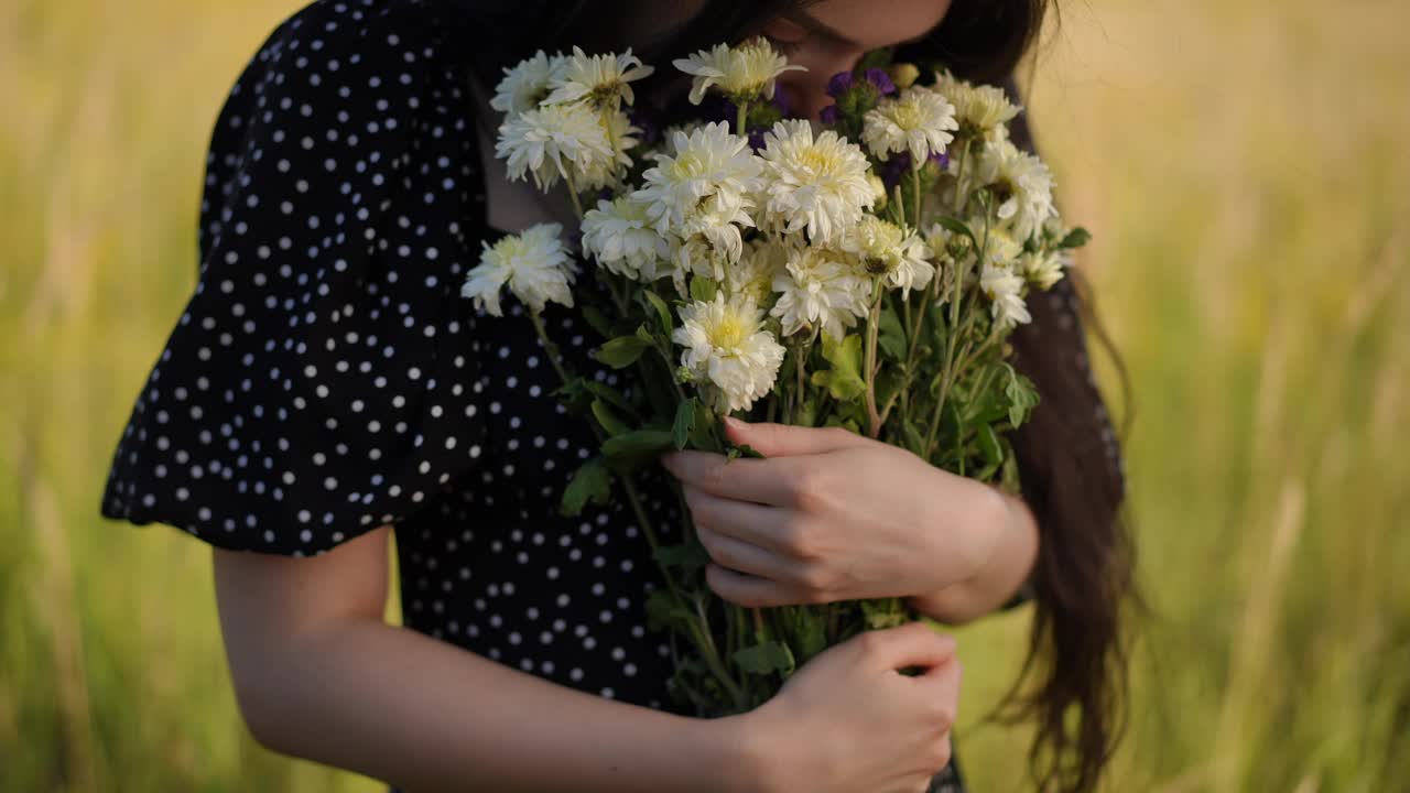 特写镜头。夏日里，一名深色长发的年轻女子站在户外，以黄绿色的草地为背景，手里拿着一大束野花左右摇摆。视频下载