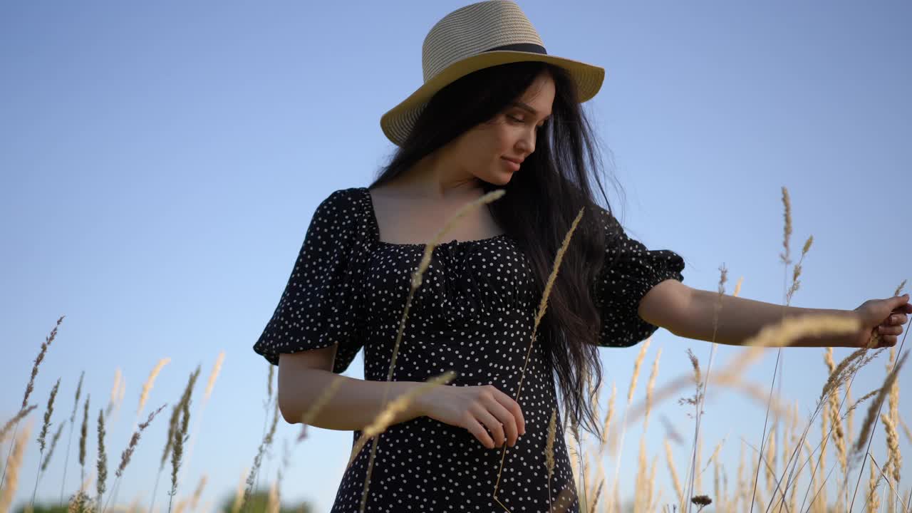 夏日，在蓝天的映衬下，站在田野里，一个黑发长发、头戴有檐帽、身穿黑色连衣裙的女孩，用手抚摸着摇曳的草地。低角。视频下载