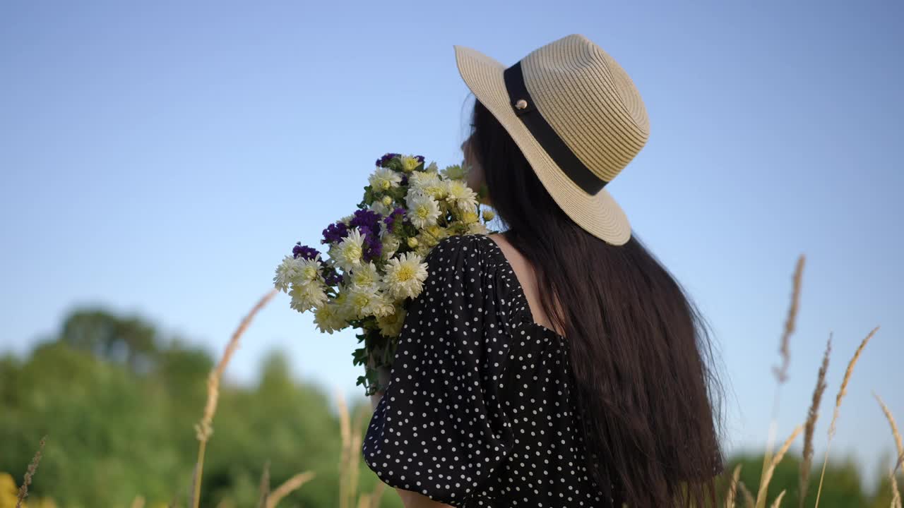 一位年轻的长发女子，头戴有檐帽，手里拿着一束野花，转向镜头。一个微笑的女孩站在夏日的田野里看着镜头。视频下载