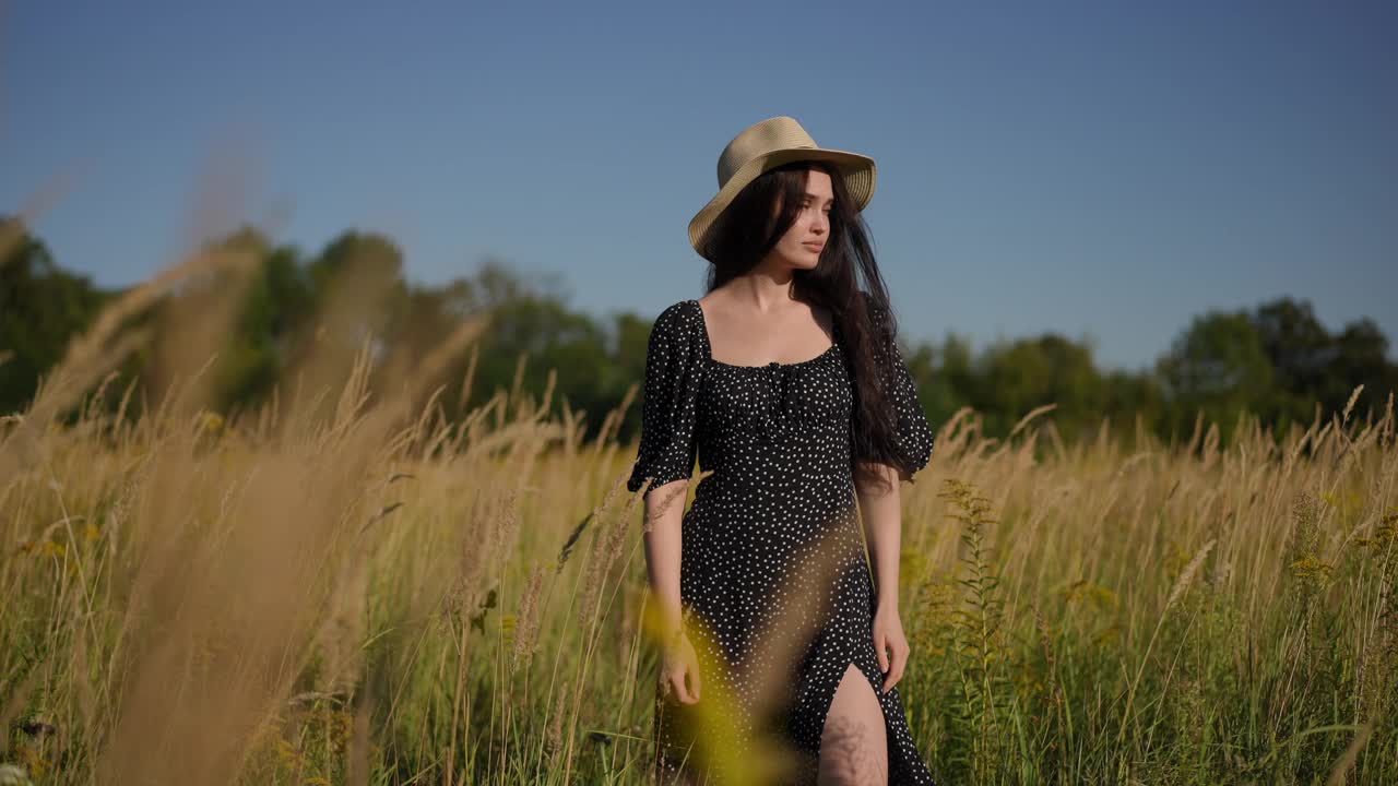 一个可爱的女孩，穿着黑色的裙子，戴着一顶有檐的帽子，站在田野里，高高的草在风中摇曳。女孩抬起头，转过身来，把帽子扔了起来，跑过田野。视频下载