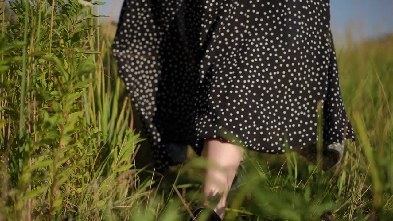 特写镜头。缓慢的运动。在一个阳光明媚的夏日，一个不知名的女人穿着黑色长裙，手里拿着一顶有檐帽，走过一片田野。身体的部分。视频下载