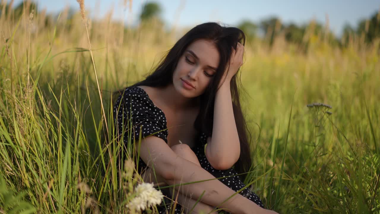 一个忧郁的黑发女孩看着白色的野花，采摘它们，闻着它们的香气，坐在一个阳光明媚的夏日田野的高绿草地上。视频下载