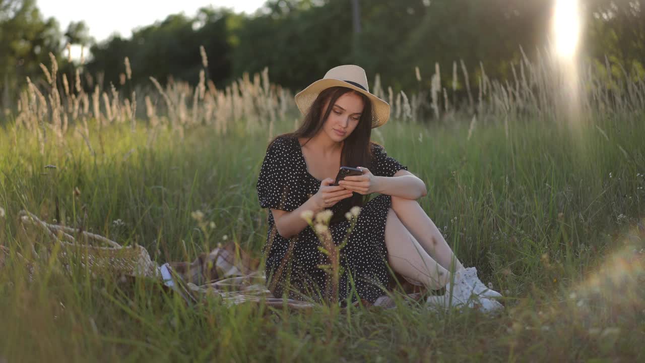 在一个阳光明媚的夏日，一位漂亮的黑发女孩戴着一顶有檐的帽子，坐在田野里高高的绿草地上，正在用手机打字。女孩笑了，看着屏幕。视频下载