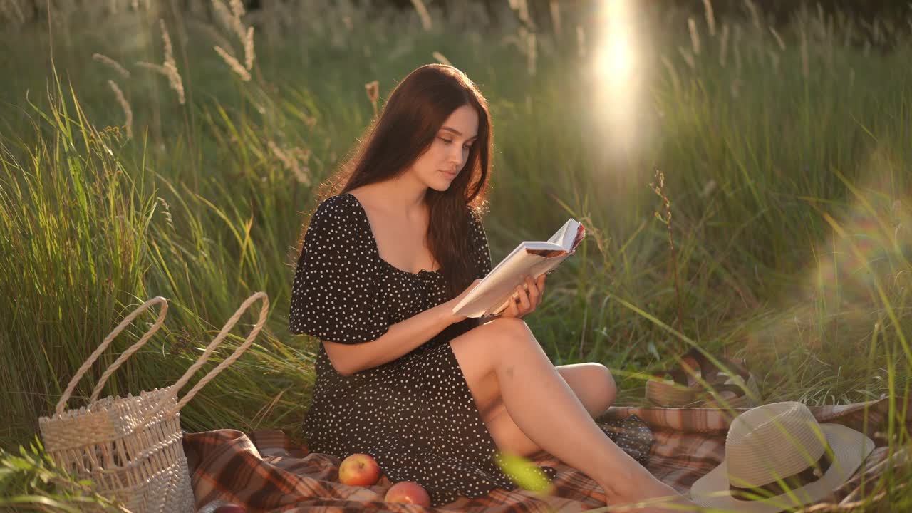 缓慢的运动。在一个阳光明媚的夏日，一个黑发长发、身穿黑色连衣裙的女孩坐在草地上读着书。在大自然中享受户外阅读的女孩。视频下载
