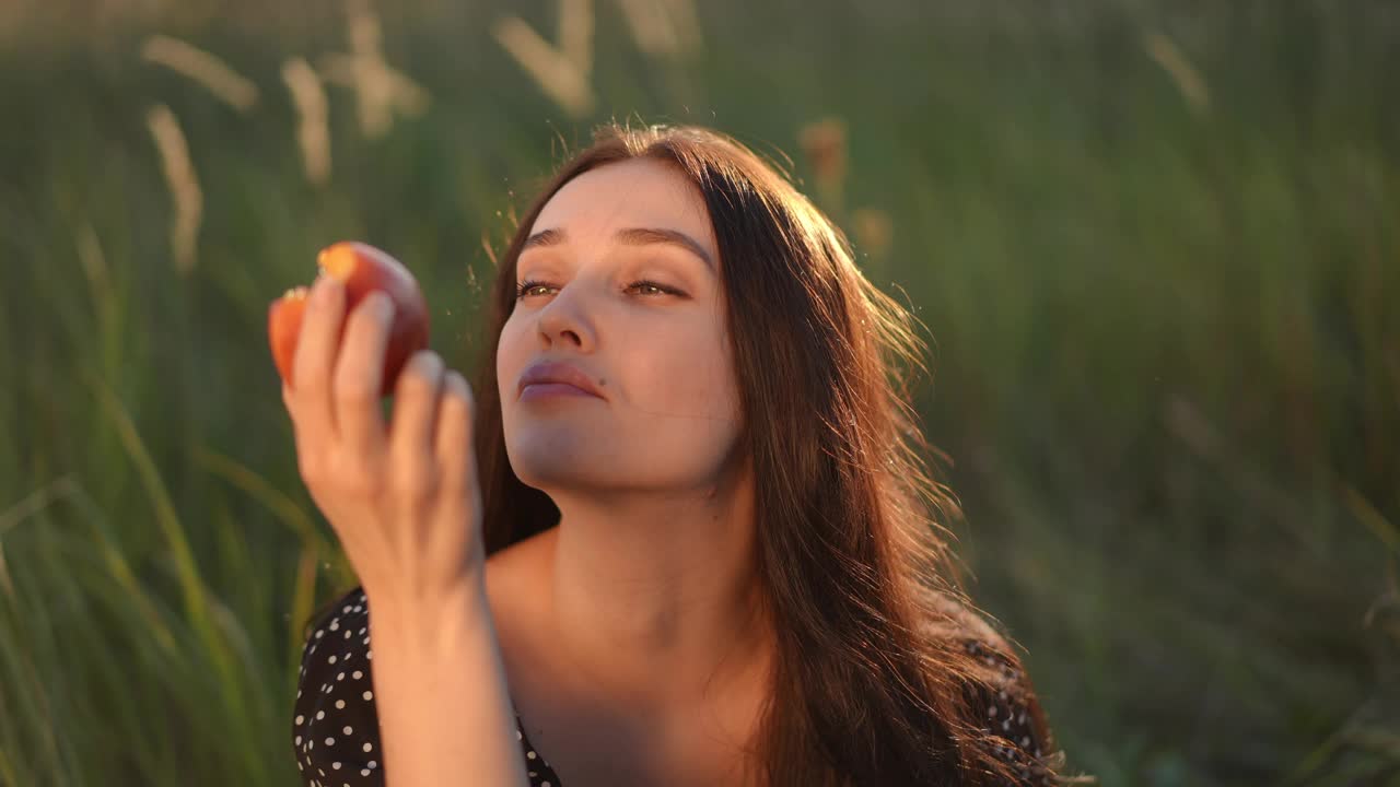 缓慢的运动。特写镜头。在大自然中野餐。在一个阳光明媚的日子里，一个戴着牙套的美丽女孩坐在田野里高高的绿草地上，吃着手里拿着的桃子，享受着它的味道。视频下载