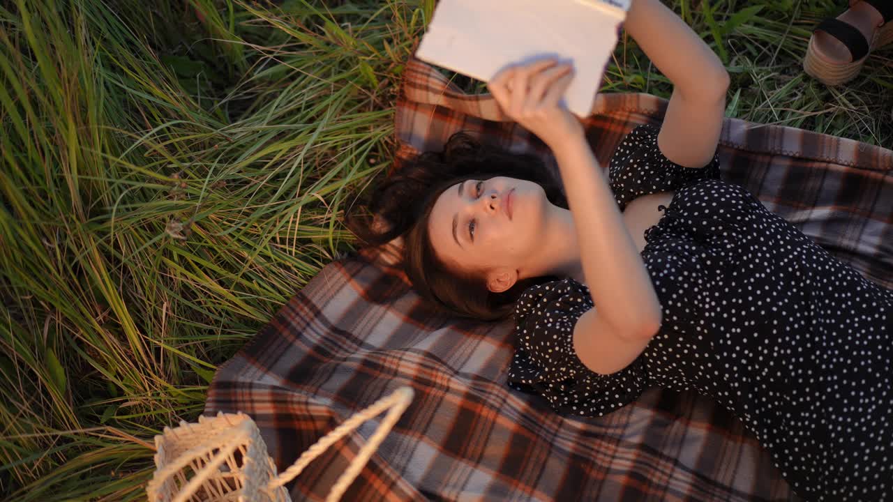 缓慢的运动。前视图。一个女孩躺在户外田野的格子上看书。黑发女人读完了书，合上了书，把它压在自己身上，闭上了眼睛。视频下载