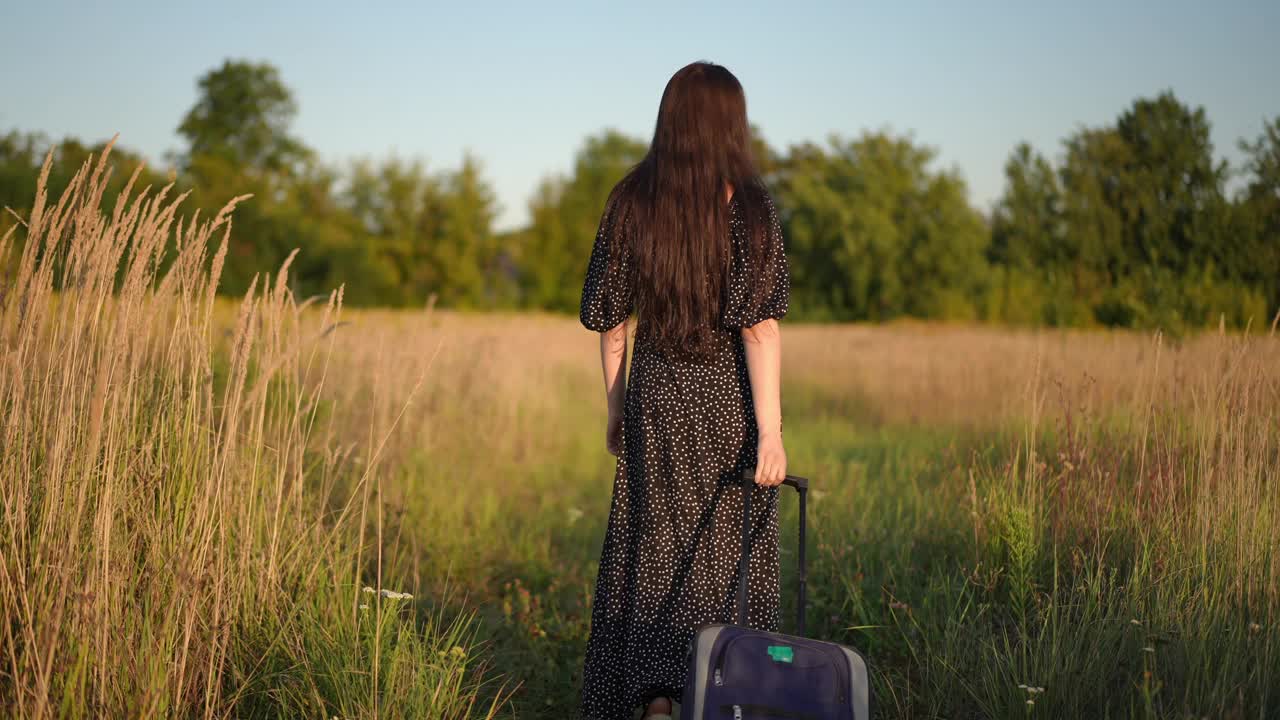 夏日夕阳下，一个黑发长发、身穿黑色连衣裙的女孩提着滚动行李箱走过田野。女孩转过身，微笑着看着镜头。视频下载