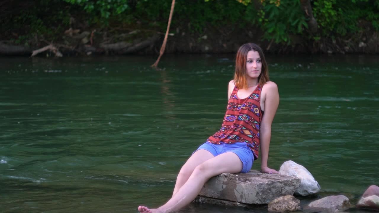 年轻女子坐在河边的石头上视频下载