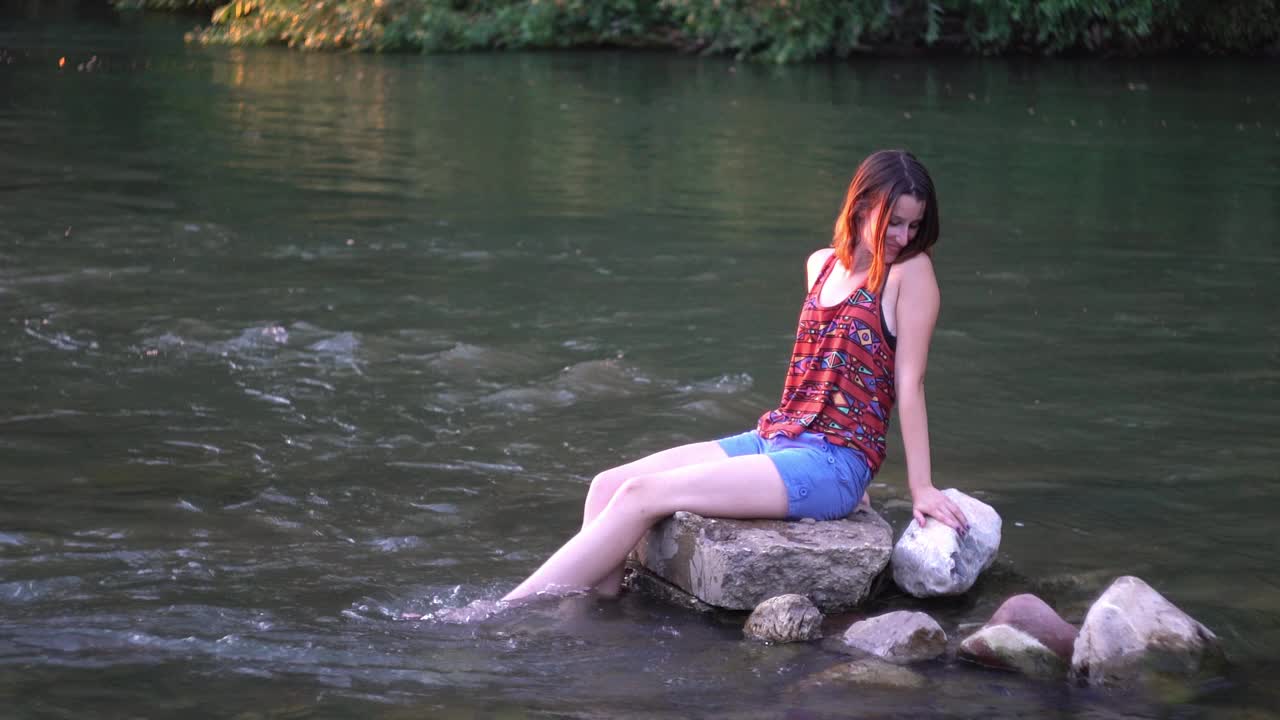 年轻女子坐在河边的石头上视频下载