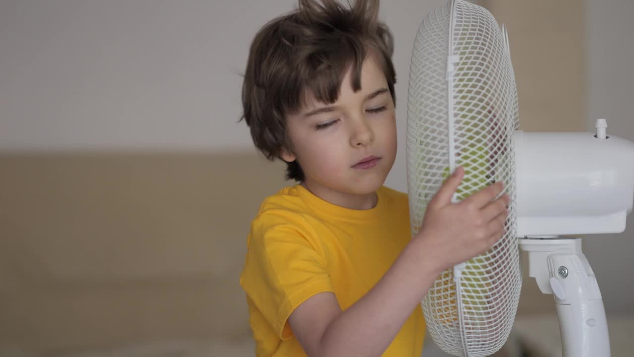 孩子在家里享受电风扇吹来的凉风。男孩在通风机前遭受高温，用电风扇冷却器给自己降温。炎热气候变化。视频下载