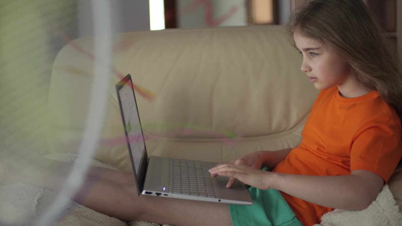孩子坐在家里的沙发上使用笔记本电脑浏览社交媒体，在家上学。酷暑的孩子享受着风扇吹来的清凉微风。大热天在家里的客厅里视频下载