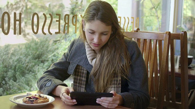 一位年轻女子独自坐在咖啡桌旁，一边用平板电脑端着一杯咖啡视频素材