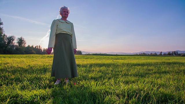 夕阳西下，老妇人站在田野上，风吹着她的裙子视频下载