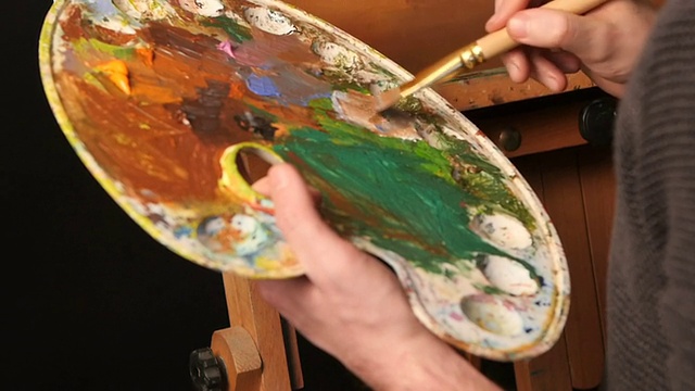 一位天才画家，深色毛衣用手中的调色板在油画上继续作画，黑色背景，背光，特写，慢动作视频下载