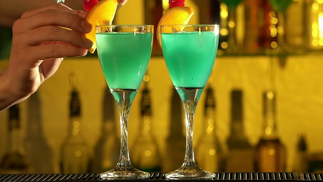 酒吧招待员将冰块放入玻璃杯，倒入三种不同的酒精液体，绿松石色，使用gayser，两个玻璃杯，用橙子和樱桃片装饰，酒吧，飞溅，慢动作视频下载