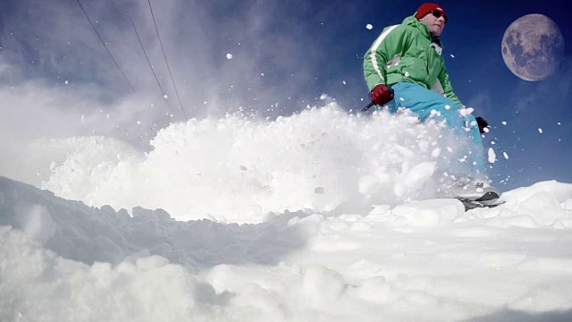 一名滑雪者在蓝天上有月亮的情况下从雪坡上滑下来的有力而充满活力的动态镜头。先是实时速度视频，然后减速来展示猛烈的雪暴。视频素材
