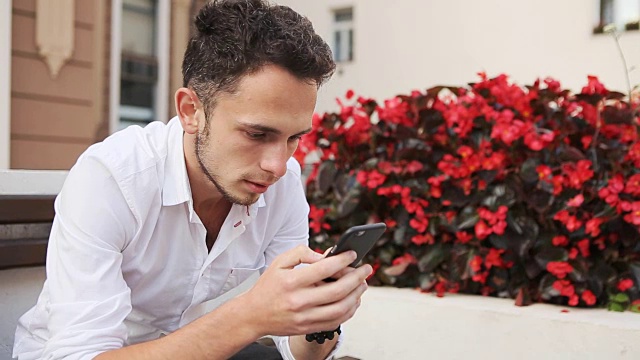 一名穿着白衬衫的年轻人坐在长凳上使用手机。在智能手机上滚动屏幕的潮人。视频下载