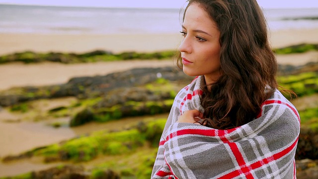 一个深思熟虑的黑发女人裹着毯子坐在海滩上视频素材