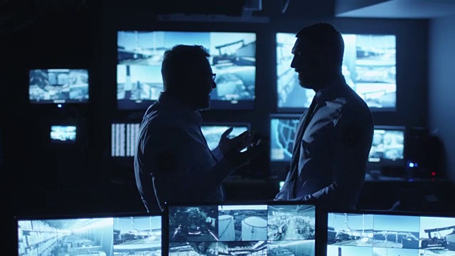 两名安全人员正在一间满是显示屏的黑暗监控室里交谈。视频素材