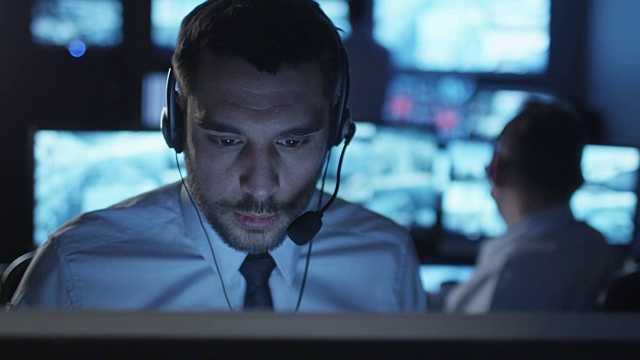 技术支持专家正在一个黑暗的充满显示屏的监理室里一边用耳机说话一边在电脑上工作。视频素材