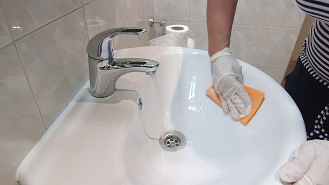 妇女戴着手套在家做家务，打扫浴室，用超细纤维布擦拭水槽和水龙头视频下载