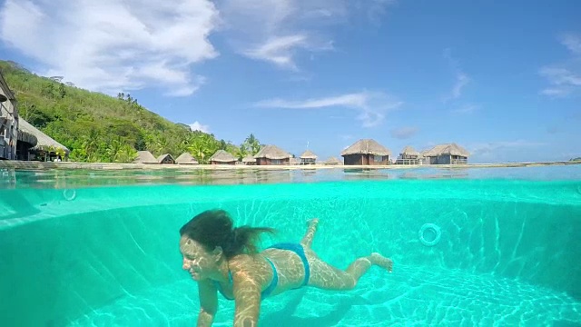 慢镜头:女子在水下游泳池游泳，背景是水上别墅视频素材