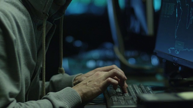 一名戴着兜帽的男性黑客在黑暗的办公室里操作电脑，电脑屏幕上显示着地图和数据。视频素材