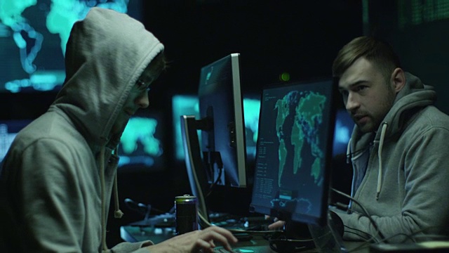 在一间昏暗的办公室里，两名穿着帽衫的黑客正在电脑上工作，电脑屏幕上显示着地图和数据。视频素材