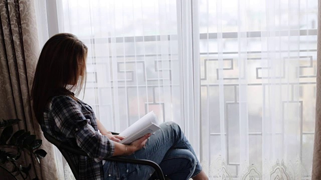 可爱的女人穿着蓝色的牛仔裤和灰色的t恤坐在窗前的桌子上，在她的客厅里看书。视频下载