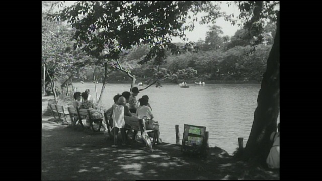 母亲和孩子们坐在公园的长椅上，观看在猪城池塘划船的人。视频下载