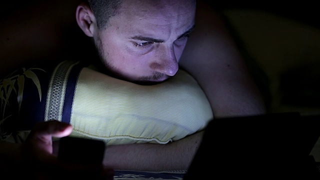 年轻人晚上在床上用平板电脑和智能手机视频下载