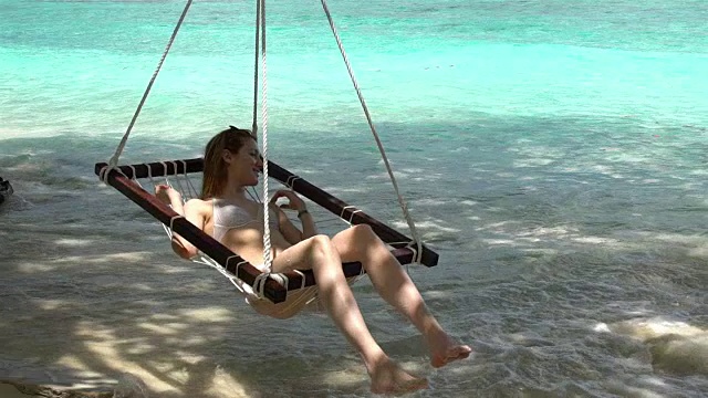 一名年轻女子在热带海滩的吊床上享受着海浪的声音。视频素材